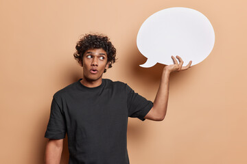 Wall Mural - Studio shot of isolated on beige background young handsome hindu guy with black curly short hair wearing dark blue tshirt holding white big speechbubble at his raised right hand and looking at it