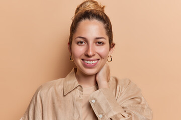 Wall Mural - Portrait of cheerful young woman keeps hand on neck smiles broadly wears shirt and golden earrings looks directly at camera isolated over brown background. People and happy emotions concept.