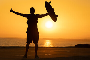 Wall Mural - Silhouette of a man showing a shaka and holding a skateboard at sunset