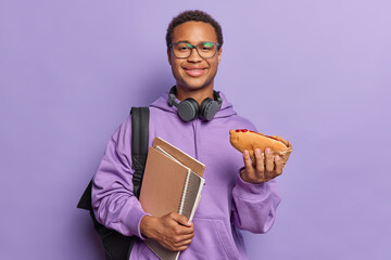 Wall Mural - Pleased hungry male student holds hot dog and notepads wants to eat dressed in casual sweatshirt wears headphones around neck and eyeglasses poses with rucksack isolated over purple background