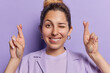Positive European woman winks eye exudes hope and optimism as she crosses her fingers winks mischievously and flashes broad smile dressed in shirt isolated over purple background. Body language