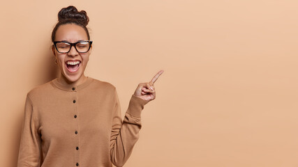 Wall Mural - Studio shot of emotional young Latin woman with hair bun points index finger on blank space for your advertising content wears spectacles and jumper poses against brown background. Look at this