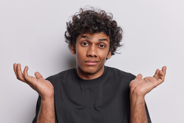 Wall Mural - Young curly haired Hindu man in his 20s wears casual black t shirt expresses sense of confusion and uncertainty spreads palms cannot answer your difficult question isolated over white background