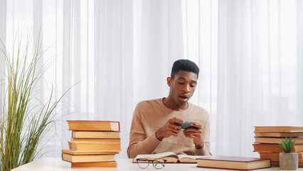 Wall Mural - Phone addiction. Gadget distraction. Study rest. Excited obsessed young man playing mobile video game relaxing at desk with books at home class.