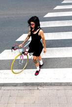 Young Ethnic Woman In Sunglasses Walking On Zebra Crossing Of Asphalt Roadway With Bicycle