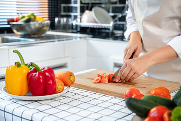 Wall Mural - Asian housewife wearing apron and using knife to slice tomato on chopping board while preparing ingredient