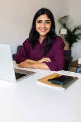 Young indian woman smile at camera working with laptop at home. Confident ethnic female sitting on living room table using computer.