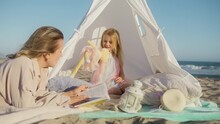 Young blonde woman creating memories for daughter. Mother lying at white tent on ocean beach and reading book to little girl, Slow motion 4K. Beautiful adorable preschooler having fun time with parent