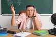 Stressed male teacher sitting at table in classroom