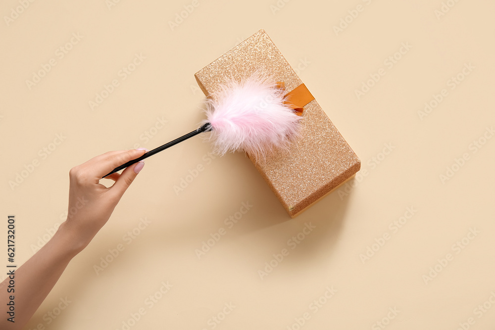 Female hand with feather stick and gift box on beige background
