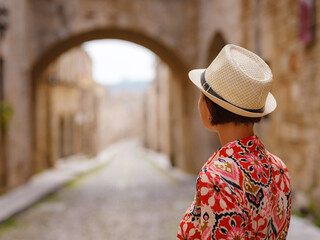 Wall Mural - summer trip to Rhodes island, Greece. Young Asian woman in ethnic red dress walks Street of Knights of Fortifications castle. female traveler visiting southern Europe. Unesco world heritage site.