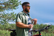 © MarijaBazarova - Cyclist have rest near the bicycle, in the forest and relax for energy in adventure workout, drinking bottle of water for hydration and  enjoying in the forest trees for wellness. Bearded man on bike