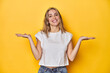 © Asier - Young blonde Caucasian woman in a white t-shirt on a yellow studio background, makes scale with arms, feels happy and confident.