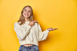 © Asier - Young blonde Caucasian woman in a white sweatshirt on a yellow studio background, excited holding a copy space on palm.
