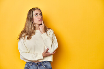 Wall Mural - Young blonde Caucasian woman in a white sweatshirt on a yellow studio background, looking sideways with doubtful and skeptical expression.