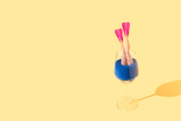 Female legs with pink flippers in blue sand sticking out of a drinking glass on a bright yellow background. Summer beach enjoyment concept.