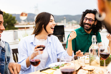 Wall Mural - Young group of friends having fun enjoying barbecue dinner party at rooftop in summer. Multiracial people reunited at home terrace laughing while celebrating together drinking red wine.