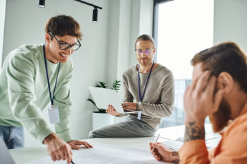 optimistic designer in eyeglasses pointing at blueprint near colleague with laptop and tattooed businessman on blurred foreground, working on startup project, collaboration in architectural studio