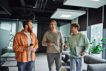 excited businessmen in eyeglasses walking with laptop and mobile phone and talking to bearded colleague in contemporary coworking space, partnership and teamwork in startup project