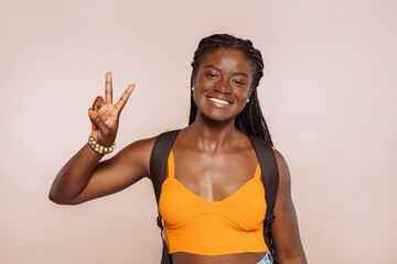 Young african female student, wearing casual orange top and backpack, gesturing peace victory sign with fingers, number two, while smiling confident and happy at studio.