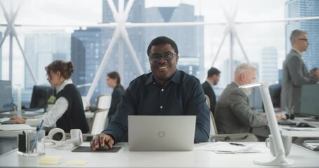 Succesful Black Man Typing On Laptop Computer In Bright Scyscraper Office With Diverse Employees In Downtown. Cheerful Male Programmer Looking At Camera And Smiling In Fintech Startup Workspace.