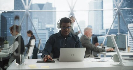 Succesful Black Man Puts On Headphones And Starts Typing On Laptop Computer In Skyscraper Office In Downtown. Professional Male Product Manager Working On a New Project In Diverse Workspace.