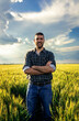 Portrait of young farmer standing in a green wheat field at sunset looking at camera.