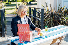 Middle-aged business woman pulls out her lunch meal at the park outdoor cafe, picnic area during her break. Takeaway balanced diet lunch box with salad. Healthy eating habits and well-being