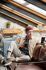 Aufkleber - Young restful businessman in casualwear texting in mobile phone and having cup of coffee while sitting on leather couch in front of camera