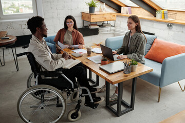 Wall Mural - Two young intercultural female managers looking at confident African American male colleague in wheelchair during discussion at meeting