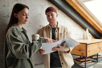 Wall Mural - Young confident businesswoman with cup of coffee pointing at paperwork with financial data while explaining it to male colleague at meeting