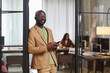 Happy young African American male designer in formalwear looking at camera while standing by open door of office and using smartphone
