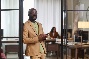 Fototapete - Happy young African American male designer in formalwear looking at camera while standing by open door of office and using smartphone