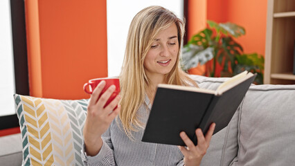 Poster - Young blonde woman reading book and drinking coffee sitting on sofa at home