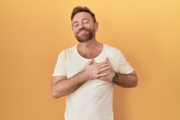 Poster - Middle age man with beard standing over yellow background smiling with hands on chest with closed eyes and grateful gesture on face. health concept.