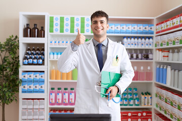 Sticker - Young hispanic man working at pharmacy drugstore holding stethoscope smiling happy and positive, thumb up doing excellent and approval sign