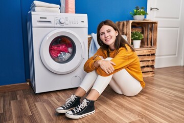 Poster - Young blonde woman sitting on floor waiting for washing machine smiling at laundry room