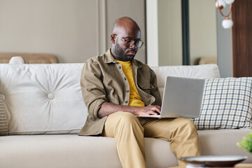 Fototapete - Serious male freelancer or creative designer sitting on couch and typing on laptop keyboard while networking or looking for online information