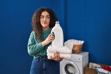 Poster - Young hispanic woman holding laundry and detergent bottle smiling looking to the side and staring away thinking.