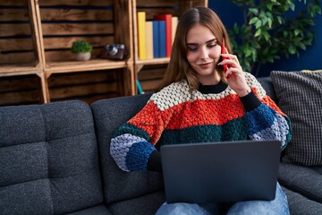 Wall Mural - Young woman using laptop and talking on smartphone at home