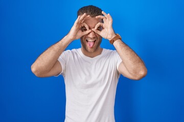 Poster - Young caucasian man standing over blue background doing ok gesture like binoculars sticking tongue out, eyes looking through fingers. crazy expression.