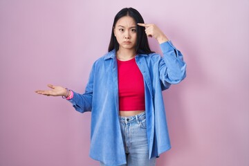 Sticker - Young asian woman standing over pink background confused and annoyed with open palm showing copy space and pointing finger to forehead. think about it.