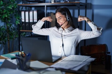 Wall Mural - Young brunette woman wearing call center agent headset working late at night showing arms muscles smiling proud. fitness concept.