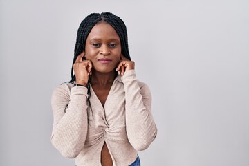 Sticker - African woman with braids standing over white background covering ears with fingers with annoyed expression for the noise of loud music. deaf concept.