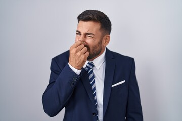 Sticker - Handsome hispanic man wearing suit and tie smelling something stinky and disgusting, intolerable smell, holding breath with fingers on nose. bad smell