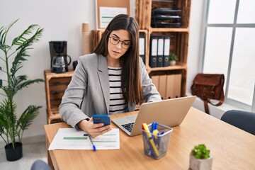 Young beautiful hispanic woman business worker using smartphone and laptop at office