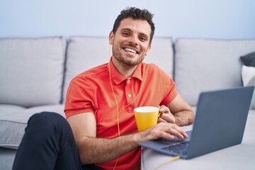 Wall Mural - Young hispanic man using laptop and drinking coffee sitting on floor at home