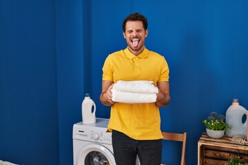 Canvas Print - Young hispanic man holding clean towels at laundry room sticking tongue out happy with funny expression.