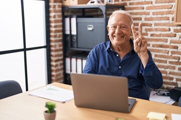 Senior man with grey hair working using computer laptop at the office smiling with happy face winking at the camera doing victory sign. number two.