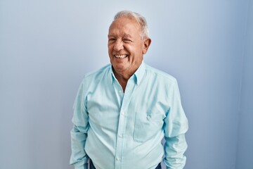 Wall Mural - Senior man with grey hair standing over blue background winking looking at the camera with sexy expression, cheerful and happy face.